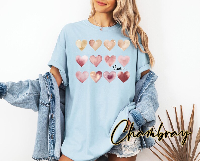 Puede incluir: Una camiseta azul claro con un dise&ntilde;o de acuarela de corazones rosas y marrones. Los corazones est&aacute;n dispuestos en una cuadr&iacute;cula y el texto "Love" est&aacute; escrito en letra negra debajo de los corazones.