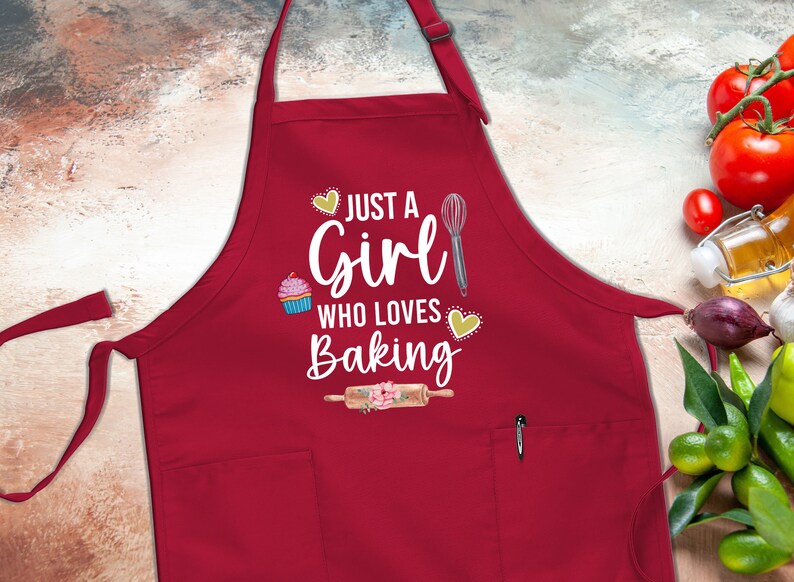 May include: Red apron with a white graphic that says "Just a Girl Who Loves Baking". The graphic includes a cupcake, a whisk, and a rolling pin.