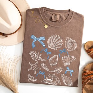 May include: A brown t-shirt featuring a seashell design, light blue bows, and a gold necklace. A beige hat with a brown band and sandals are also in the image. The t-shirt has the size XL-TG-EG printed on the tag.