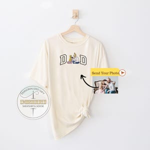 May include: Cream-colored t-shirt with the word "DAD" in a collegiate-style font. The graphic features a family illustration. A small photo is attached, with the text "Send Your Photo". The shirt is hanging on a wooden hanger.
