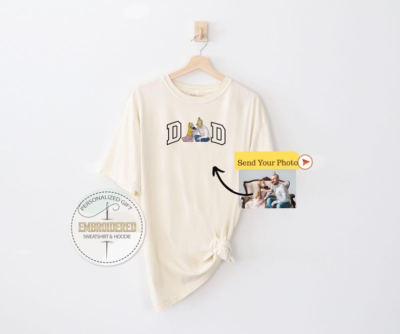 May include: Cream-colored t-shirt with the word "DAD" in a collegiate-style font. The graphic features a family illustration. A small photo is attached, with the text "Send Your Photo". The shirt is hanging on a wooden hanger.