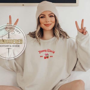 May include: A sand-colored sweatshirt with the embroidered text "Disney World Est. 1971" in red. The sweatshirt has a crew neck and long sleeves. A beige beanie is worn on the head. The image also includes a personalized gift tag.