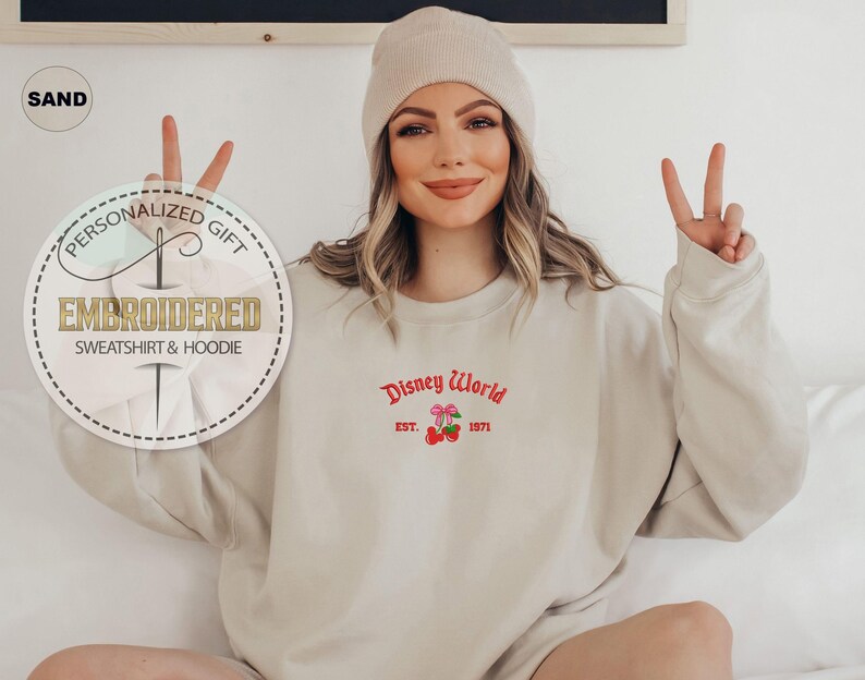 May include: A sand-colored sweatshirt with the embroidered text "Disney World Est. 1971" in red. The sweatshirt has a crew neck and long sleeves. A beige beanie is worn on the head. The image also includes a personalized gift tag.