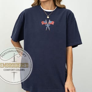 May include: Navy blue t-shirt with an embroidered design of two American flags crossed at the stems. The shirt has short sleeves and a crew neck. The text "EMBROIDERED COMFORT COLORS" is printed on a circular label.