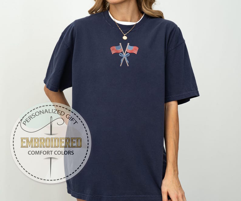 May include: Navy blue t-shirt with an embroidered design of two American flags crossed at the stems. The shirt has short sleeves and a crew neck. The text "EMBROIDERED COMFORT COLORS" is printed on a circular label.