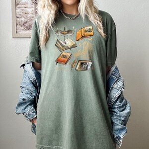 May include: Olive green t-shirt with a fantasy book graphic. The design features open books, a dragon, and the text "Iron Flame", "Fourth Wing", and "Onyx Storm". The shirt has a relaxed fit.