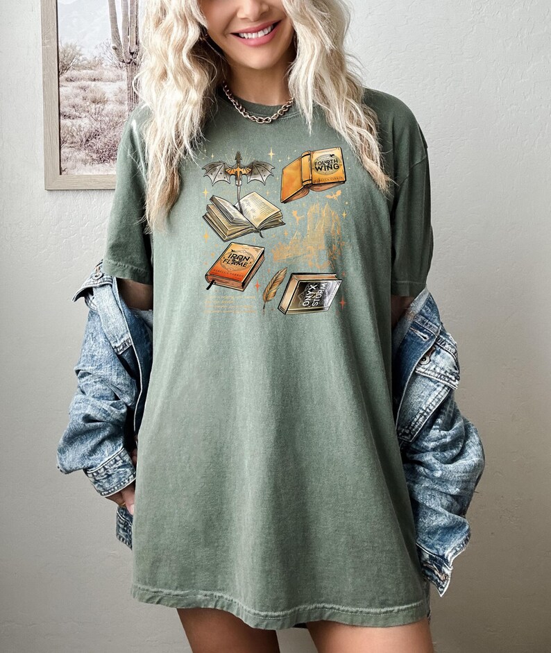 May include: Olive green t-shirt with a fantasy book graphic. The design features open books, a dragon, and the text "Iron Flame", "Fourth Wing", and "Onyx Storm". The shirt has a relaxed fit.