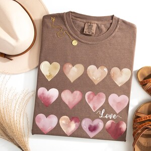 Puede incluir: Una camiseta marr&oacute;n con un dise&ntilde;o de acuarela de corazones rosas y beige. La camiseta tiene la palabra "Love" escrita en cursiva en la parte inferior.
