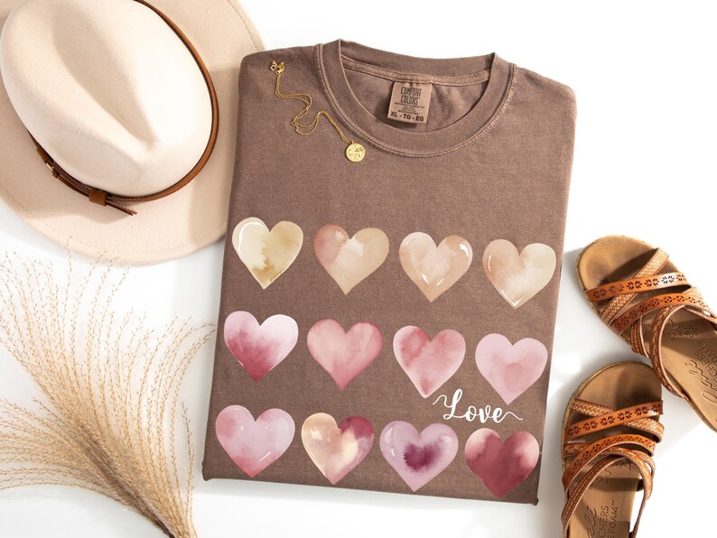 Puede incluir: Una camiseta marr&oacute;n con un dise&ntilde;o de acuarela de corazones rosas y beige. La camiseta tiene la palabra "Love" escrita en cursiva en la parte inferior.