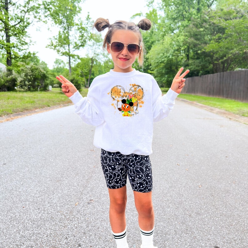May include: A young person wearing a white sweatshirt with a Mickey Mouse design, black biker shorts with a smiley face pattern, sunglasses, and white socks with black stripes. The person is standing on a road and giving a peace sign.
