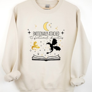 May include: A cream-colored sweatshirt with a black and gold graphic design. The graphic features a crescent moon, stars, a book, and two dragons. The text on the sweatshirt reads "Emotionally attached to fictional characters".