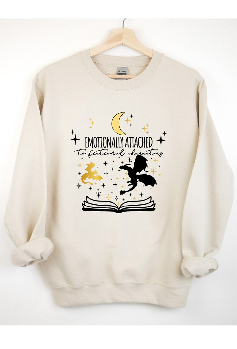 May include: A cream-colored sweatshirt with a black and gold graphic design. The graphic features a crescent moon, stars, a book, and two dragons. The text on the sweatshirt reads "Emotionally attached to fictional characters".