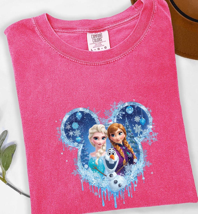 May include: A pink Comfort Colors t-shirt featuring a graphic of Elsa, Anna, and Olaf from Frozen, set within a Mickey Mouse-shaped design with snowflakes and blue accents. The shirt has a crew neck and a tag.