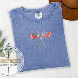 May include: A blue Comfort Colors t-shirt with an embroidered design of two crossed American flags. The flags are red and white striped with blue fields and are tied together with a blue bow. The shirt has a crew neck and a tag that says "Comfort Colors".