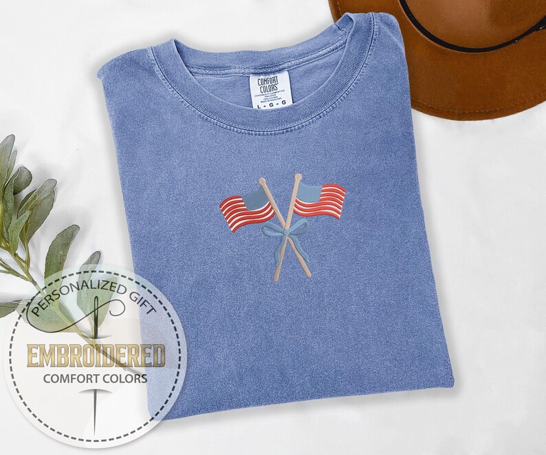 May include: A blue Comfort Colors t-shirt with an embroidered design of two crossed American flags. The flags are red and white striped with blue fields and are tied together with a blue bow. The shirt has a crew neck and a tag that says "Comfort Colors".