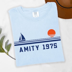 May include: Light blue t-shirt with a sailboat and sunset graphic. The design includes the text "AMITY 1975" in navy blue. The shirt is a Comfort Colors brand.