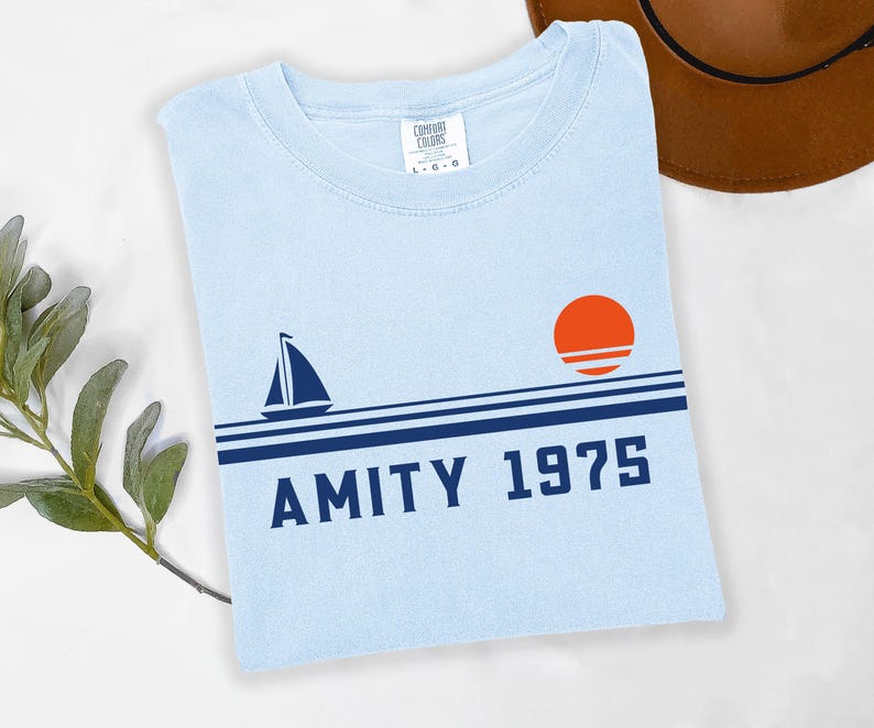 May include: Light blue t-shirt with a sailboat and sunset graphic. The design includes the text "AMITY 1975" in navy blue. The shirt is a Comfort Colors brand.