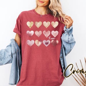 Puede incluir: Una camiseta roja con un dise&ntilde;o de acuarela de corazones rosas y dorados. Los corazones est&aacute;n dispuestos en una fila y la palabra "Love" est&aacute; escrita en blanco debajo de los corazones.