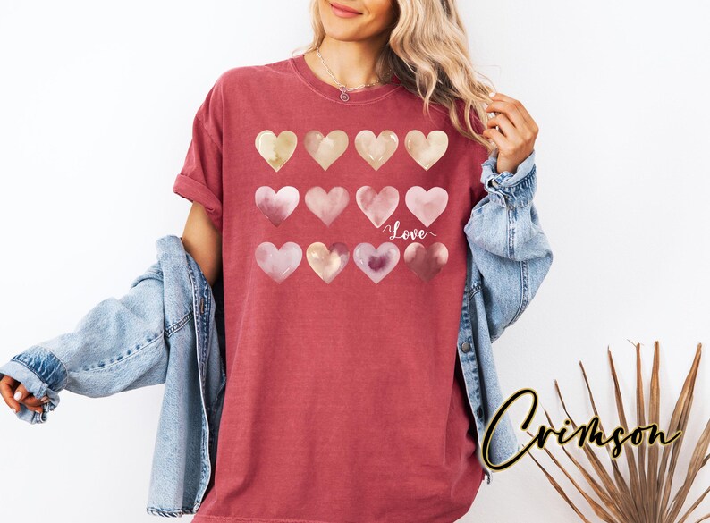Puede incluir: Una camiseta roja con un dise&ntilde;o de acuarela de corazones rosas y dorados. Los corazones est&aacute;n dispuestos en una fila y la palabra "Love" est&aacute; escrita en blanco debajo de los corazones.
