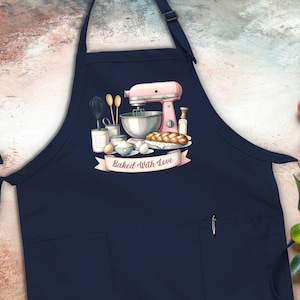 May include: A navy blue apron with a large pocket and adjustable straps. The apron features a pink stand mixer, bread, and other baking supplies with the text "Baked With Love".