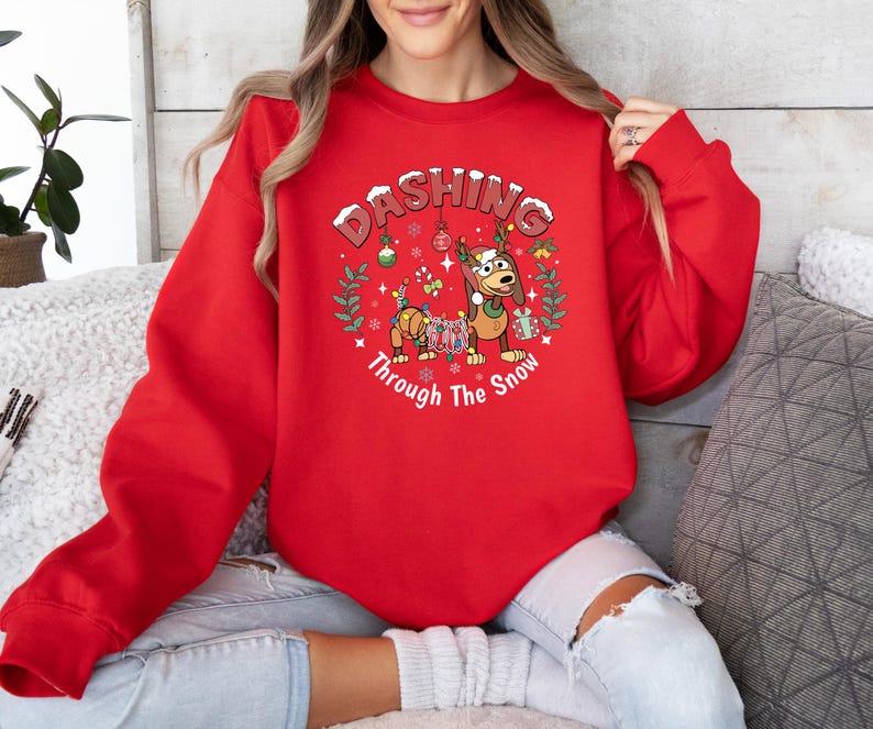 May include: A red sweatshirt with the text "Dashing Through The Snow" and a cartoon graphic of a dog wearing reindeer antlers and a dachshund dog decorated with Christmas lights. The sweatshirt is a long-sleeved crew neck.