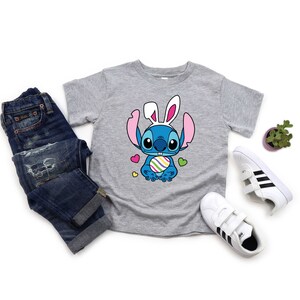 May include: A gray t-shirt with a blue cartoon character wearing bunny ears and holding a colorful Easter egg. The character is surrounded by pink hearts. The shirt is laid out on a white surface with a pair of blue jeans, a potted plant, and two pairs of white sneakers with black stripes.