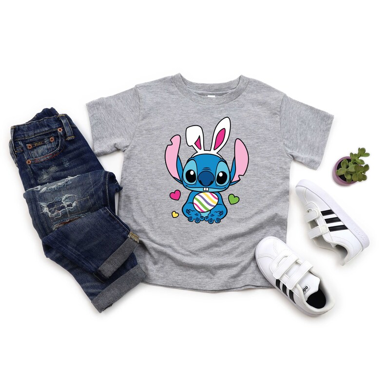 May include: A gray t-shirt with a blue cartoon character wearing bunny ears and holding a colorful Easter egg. The character is surrounded by pink hearts. The shirt is laid out on a white surface with a pair of blue jeans, a potted plant, and two pairs of white sneakers with black stripes.