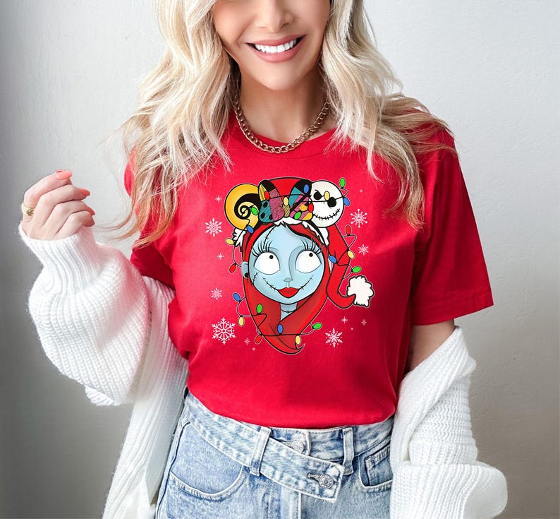 Puede incluir: Camiseta roja con un gr&aacute;fico navide&ntilde;o con Sally de Pesadilla antes de Navidad. Sally lleva un gorro de Pap&aacute; Noel y tiene luces de Navidad envueltas a su alrededor. La cabeza de Jack Skellington est&aacute; encima de su cabeza.