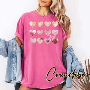 Puede incluir: Una camiseta rosa con un dise&ntilde;o de varios corazones de acuarela en tonos rosa y dorado. Los corazones est&aacute;n dispuestos en filas y la palabra "Love" est&aacute; escrita en blanco debajo de los corazones.