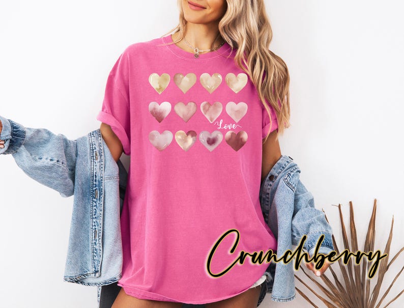 Puede incluir: Una camiseta rosa con un dise&ntilde;o de varios corazones de acuarela en tonos rosa y dorado. Los corazones est&aacute;n dispuestos en filas y la palabra "Love" est&aacute; escrita en blanco debajo de los corazones.