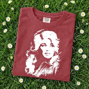 May include: A brick red short-sleeved t-shirt featuring a white graphic print of a woman with voluminous hair. The inner neck label displays the text Comfort Colors. The shirt is made of cotton and has a crew neckline and a standard fit design.