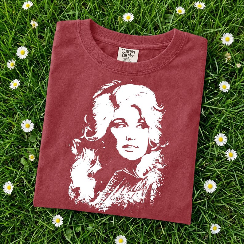 May include: A brick red short-sleeved t-shirt featuring a white graphic print of a woman with voluminous hair. The inner neck label displays the text Comfort Colors. The shirt is made of cotton and has a crew neckline and a standard fit design.