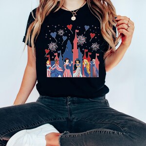 May include: Black t-shirt featuring a patriotic design with a castle silhouette and fireworks. The graphic includes several princesses in red, white, and blue attire. The design is festive and suitable for Independence Day celebrations.