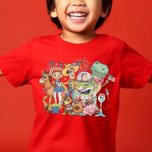 Puede incluir: Una camiseta roja de manga corta con un estampado gr&aacute;fico colorido de varios personajes de Toy Story, incluidos Woody, Buzz Lightyear, Jessie, Rex y Forky, todos decorados con accesorios patri&oacute;ticos con la bandera estadounidense y fuegos artificiales.
