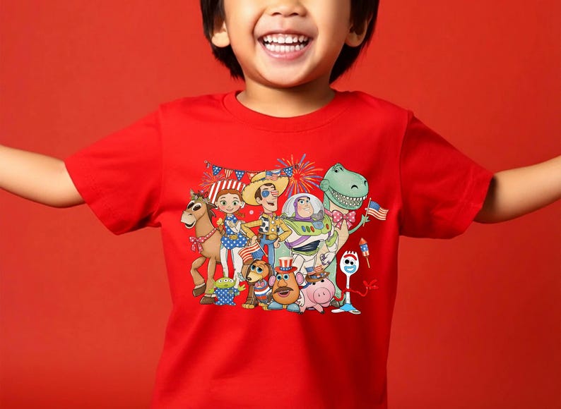 Puede incluir: Una camiseta roja de manga corta con un estampado gr&aacute;fico colorido de varios personajes de Toy Story, incluidos Woody, Buzz Lightyear, Jessie, Rex y Forky, todos decorados con accesorios patri&oacute;ticos con la bandera estadounidense y fuegos artificiales.