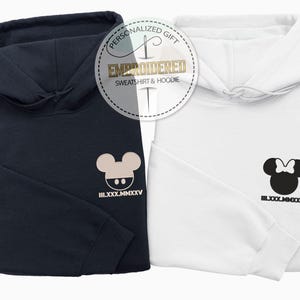 May include: Two folded hoodies, one navy blue and one white, are displayed together. Each hoodie has a cartoon mouse head design on the front. The navy hoodie has a beige mouse head, while the white hoodie has a black mouse head with a bow.
