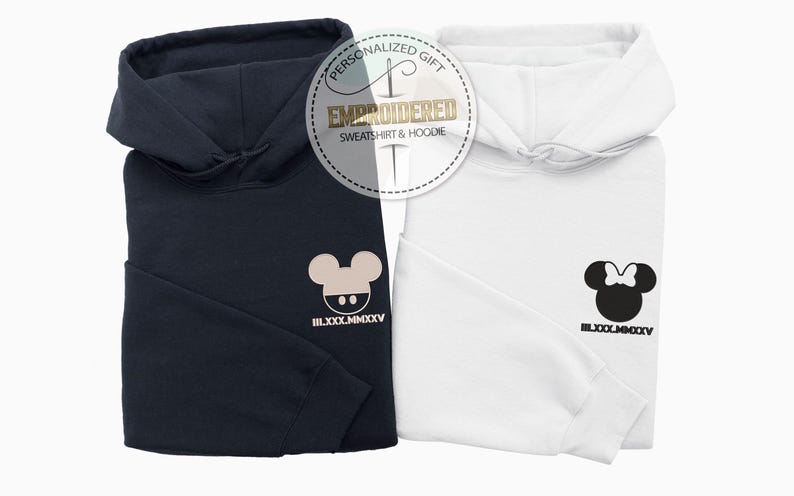 May include: Two folded hoodies, one navy blue and one white, are displayed together. Each hoodie has a cartoon mouse head design on the front. The navy hoodie has a beige mouse head, while the white hoodie has a black mouse head with a bow.