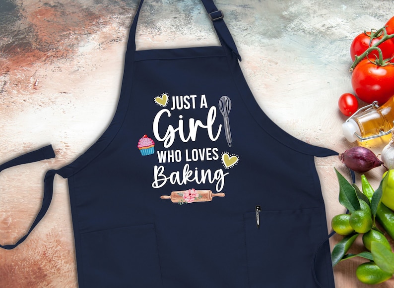 May include: A navy blue apron with a white graphic that says "Just a Girl Who Loves Baking". The graphic includes a cupcake, a whisk, a rolling pin, and hearts.