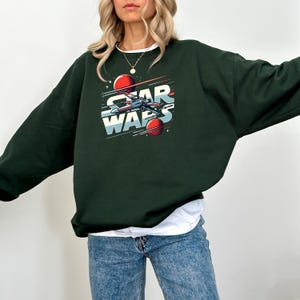 Puede incluir: Una sudadera de color verde oscuro con un estampado gr&aacute;fico de una nave espacial y el texto "Star Wars" en letras blancas y rojas.