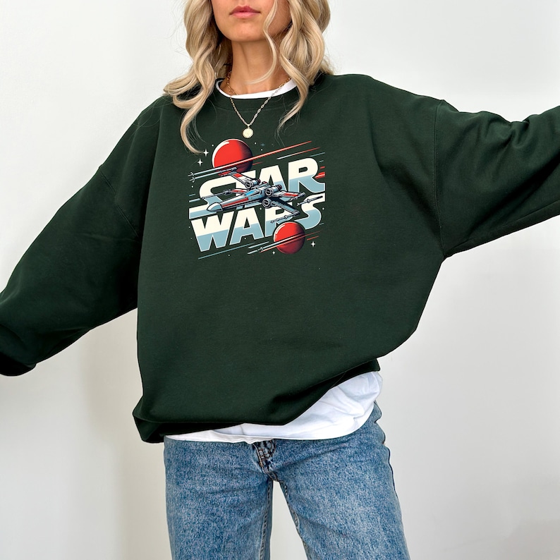 Puede incluir: Una sudadera de color verde oscuro con un estampado gr&aacute;fico de una nave espacial y el texto "Star Wars" en letras blancas y rojas.