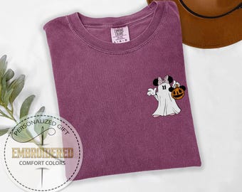 May include: A purple Comfort Colors t-shirt with a Halloween-themed embroidered design. The design features a cartoon ghost with Minnie Mouse ears holding a jack-o'-lantern. The shirt has a crew neck and short sleeves. The text "EMBROIDERED COMFORT COLORS" is also visible.