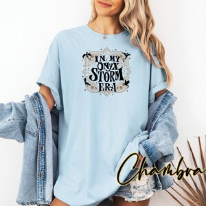 May include: A light blue t-shirt with a black graphic design that says "In My Onyx Storm Era". The graphic design features a pentagram and stylized dragons.