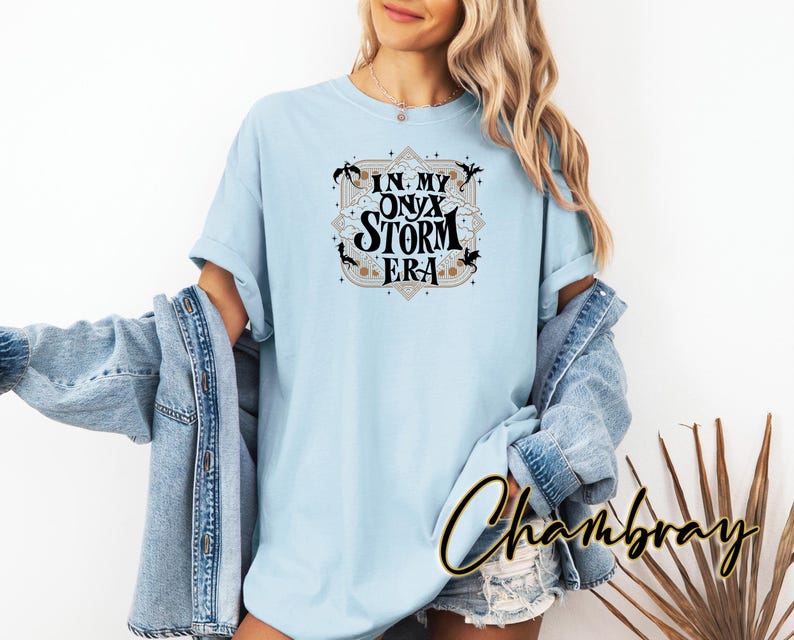 May include: A light blue t-shirt with a black graphic design that says "In My Onyx Storm Era". The graphic design features a pentagram and stylized dragons.