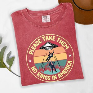 Op de afbeelding: Rood Comfort Colors T-shirt met een cirkelvormige afbeelding. De afbeelding toont een UFO die twee figuren ontvoert, met de tekst "PLEASE TAKE THEM NO KINGS IN AMERICA" in een gele cirkel. Het shirt heeft een ronde hals en korte mouwen.