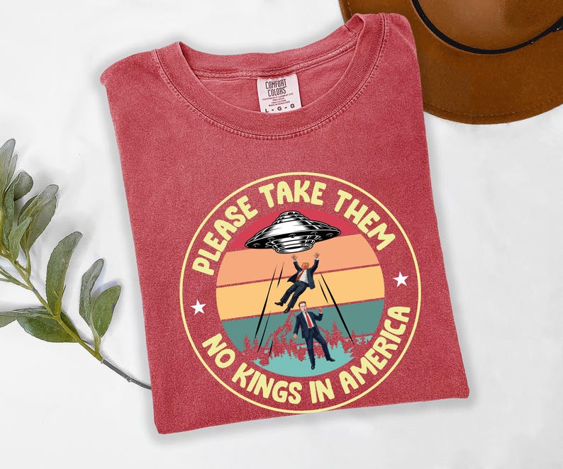 Op de afbeelding: Rood Comfort Colors T-shirt met een cirkelvormige afbeelding. De afbeelding toont een UFO die twee figuren ontvoert, met de tekst "PLEASE TAKE THEM NO KINGS IN AMERICA" in een gele cirkel. Het shirt heeft een ronde hals en korte mouwen.
