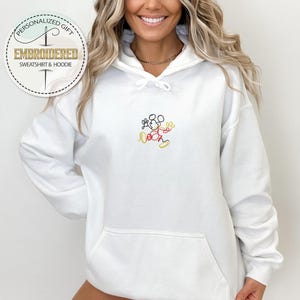 May include: White hoodie with a black and red embroidered Mickey Mouse design. The hoodie has a drawstring hood and a kangaroo pocket.