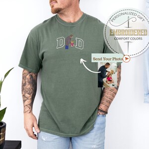 May include: Olive green t-shirt with embroidered "DAD" lettering and a graphic of a father and child. The shirt features a small photo insert and the text "PERSONALIZED GIFT EMBROIDERED COMFORT COLORS".