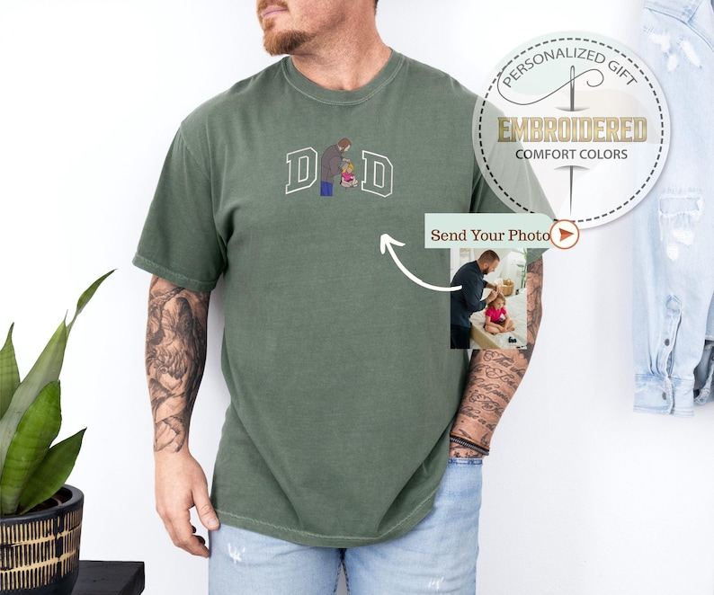 May include: Olive green t-shirt with embroidered "DAD" lettering and a graphic of a father and child. The shirt features a small photo insert and the text "PERSONALIZED GIFT EMBROIDERED COMFORT COLORS".