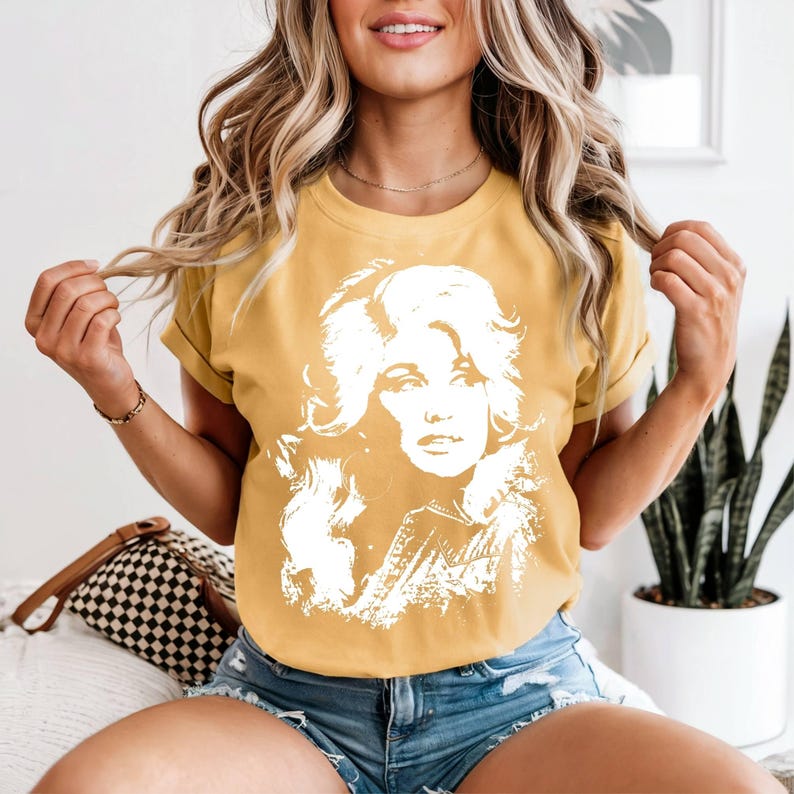 May include: A mustard yellow short-sleeved t-shirt featuring a large, white, high-contrast graphic print of a woman with voluminous hair. The shirt has a crew neckline and a relaxed fit, showcasing a detailed portrait illustration on the front.