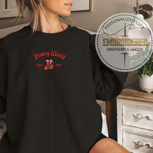 May include: Black sweatshirt with embroidered "Disney World" text in red, along with a cherry and bow design. The text "EST. 1971" is also included. The sweatshirt is a personalized gift.